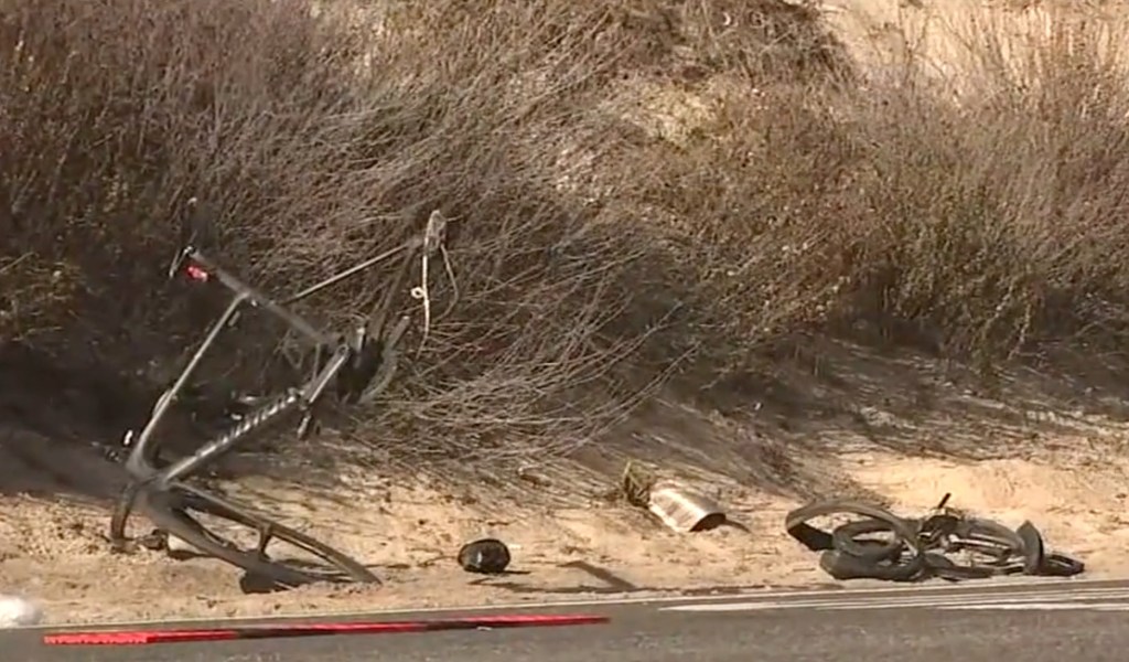 Debris, including a mangled bicycle, on the shoulder of a road next to a sandy bank with dry brush.