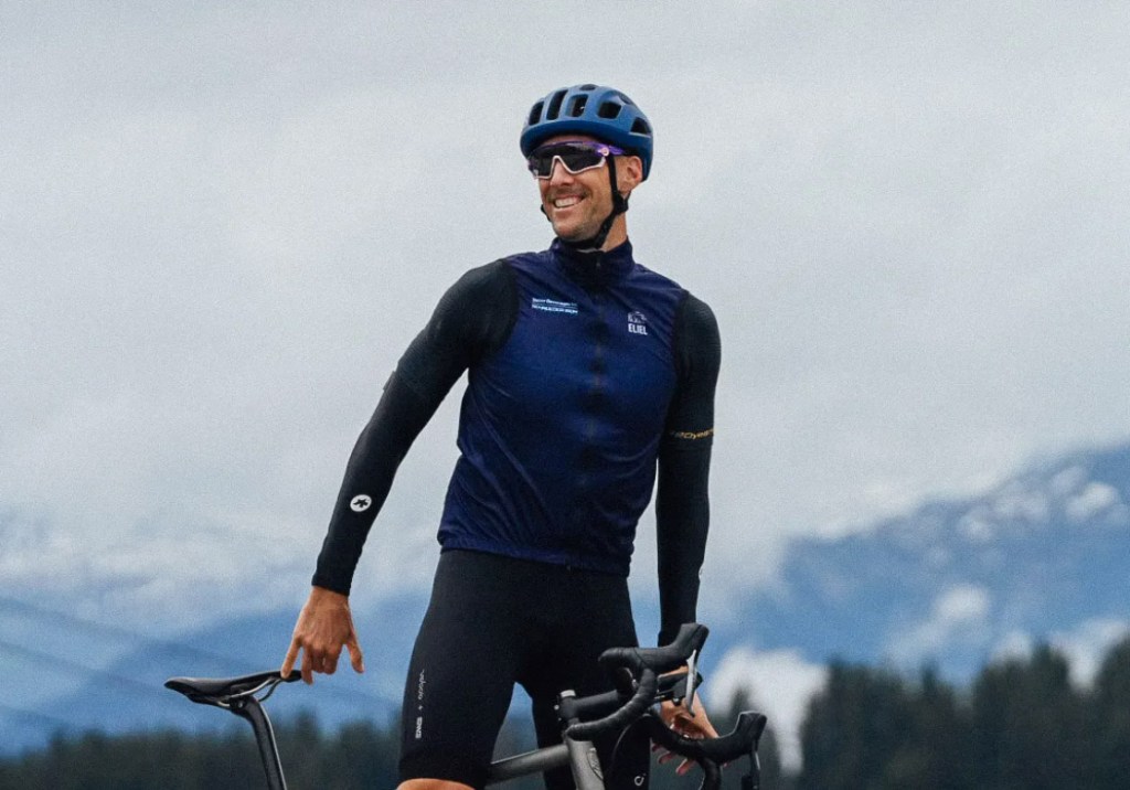 A man in cycling gear and helmet smiling while holding a bicycle.