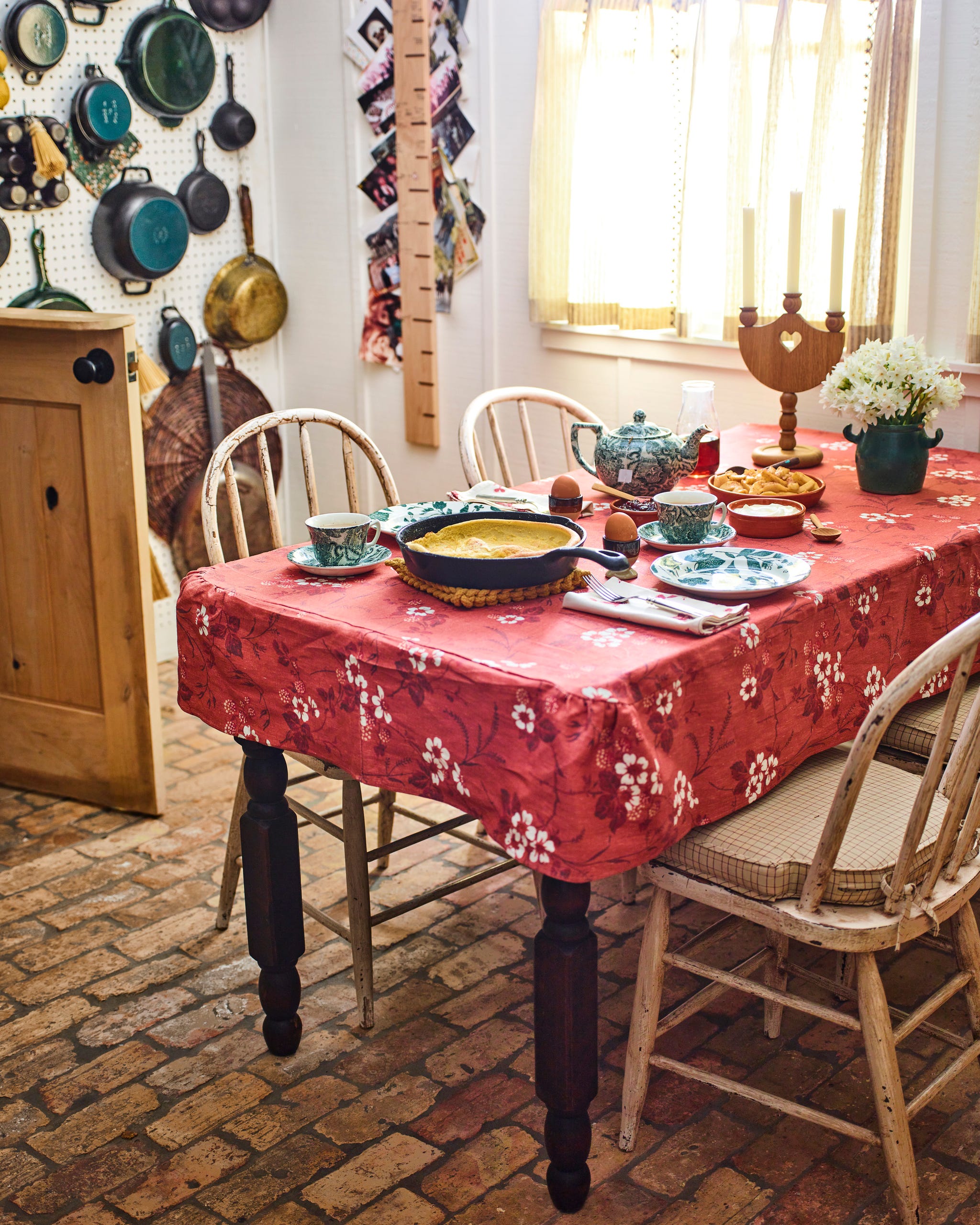kitchen table with red floral tablecloth and wood chairs around it and a collection of pots hanging on the wall behind it