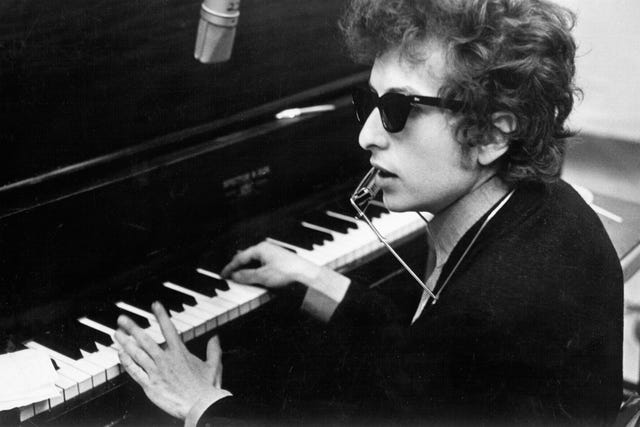 black and white photo of bob dylan at a piano, wearing sunglasses and singing into a vintage microphone—captioned with his quote: “he not busy being born is busy dying.”