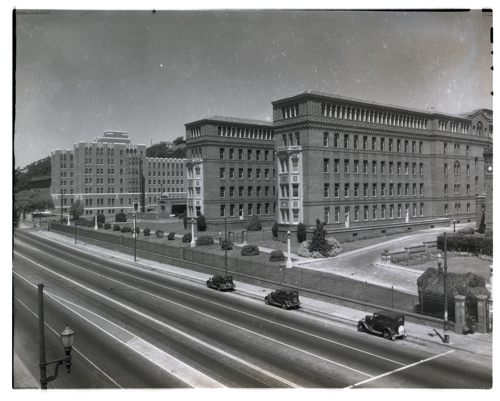 Black-and-white photo of large brick institutional buildings beside a wide, empty road with a few vintage cars parked along the curb.