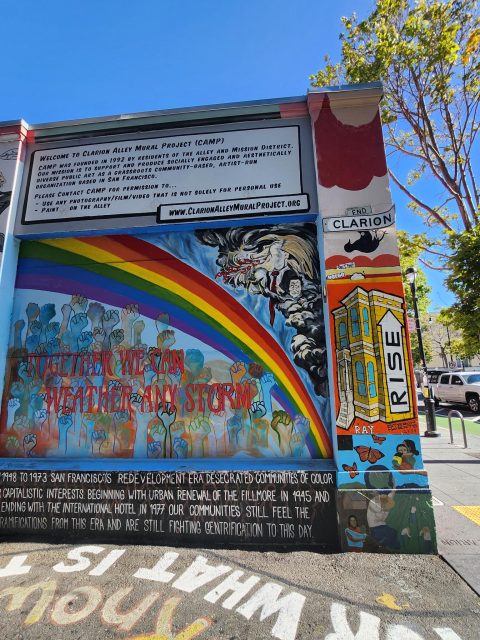 A colorful mural on a city wall features a rainbow, people, and a tiger, with the text "Together We Can Weather Any Storm." Adjacent panels display "Clarion" and "Rise" with building art.