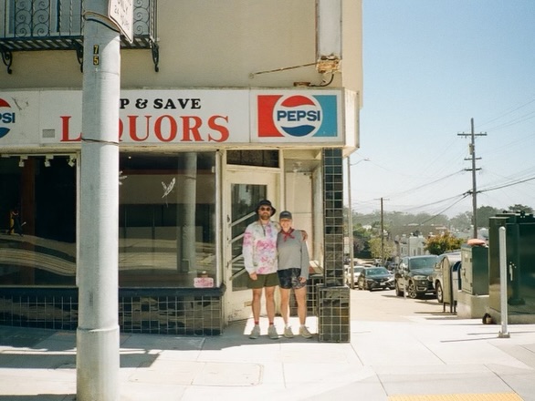 Two people stand on a sunny street corner in front of a liquor store with a large Pepsi sign. Cars are parked along the street in the background.