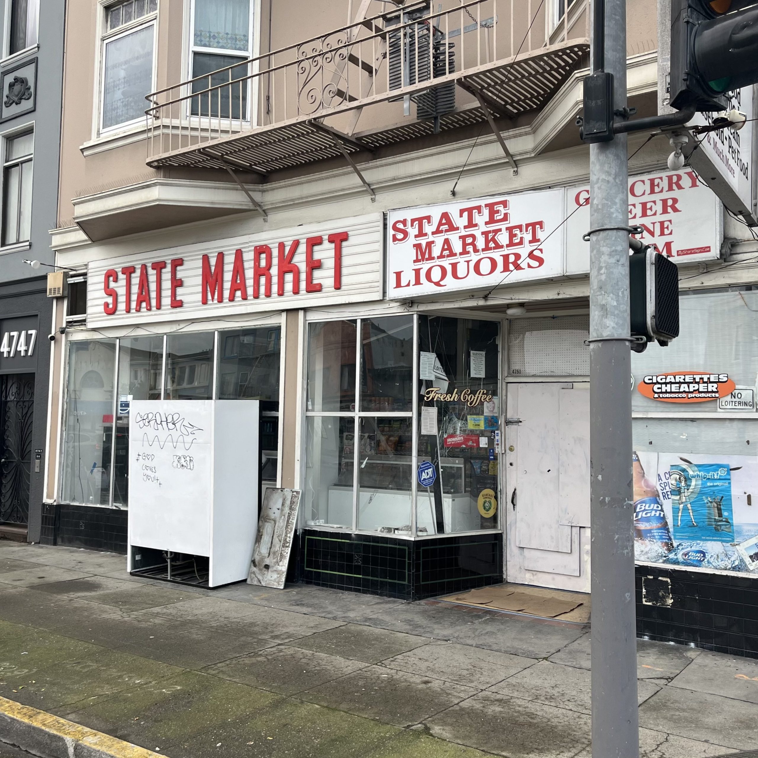Street view of a corner store called “State Market & Liquors” with signs for groceries, beer, and cigarettes; storefront appears worn and windows display various posters and ads.