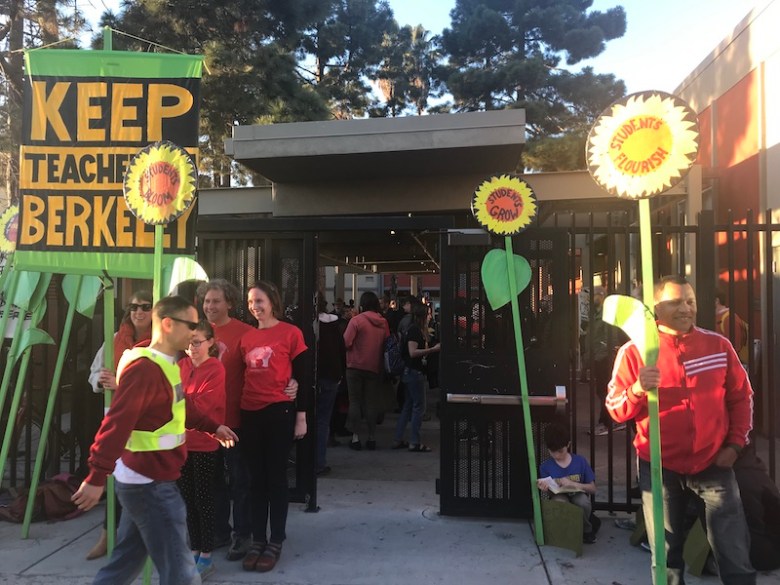 Several red-clad people stand outside the gate to a courtyard, holding tall signs shaped like sunflowers. One has a huge sign that says, "Keep teachers in Berkeley."