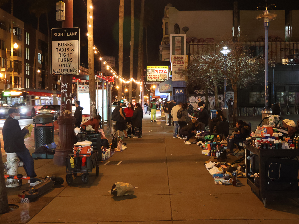 A nighttime street scene with people gathered along the sidewalk selling various items under illuminated string lights. A bus is visible in the background, and storefronts are lit up.