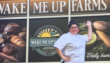 a woman pointing to a wall art sign that says wake me up farms and features loaves of bread