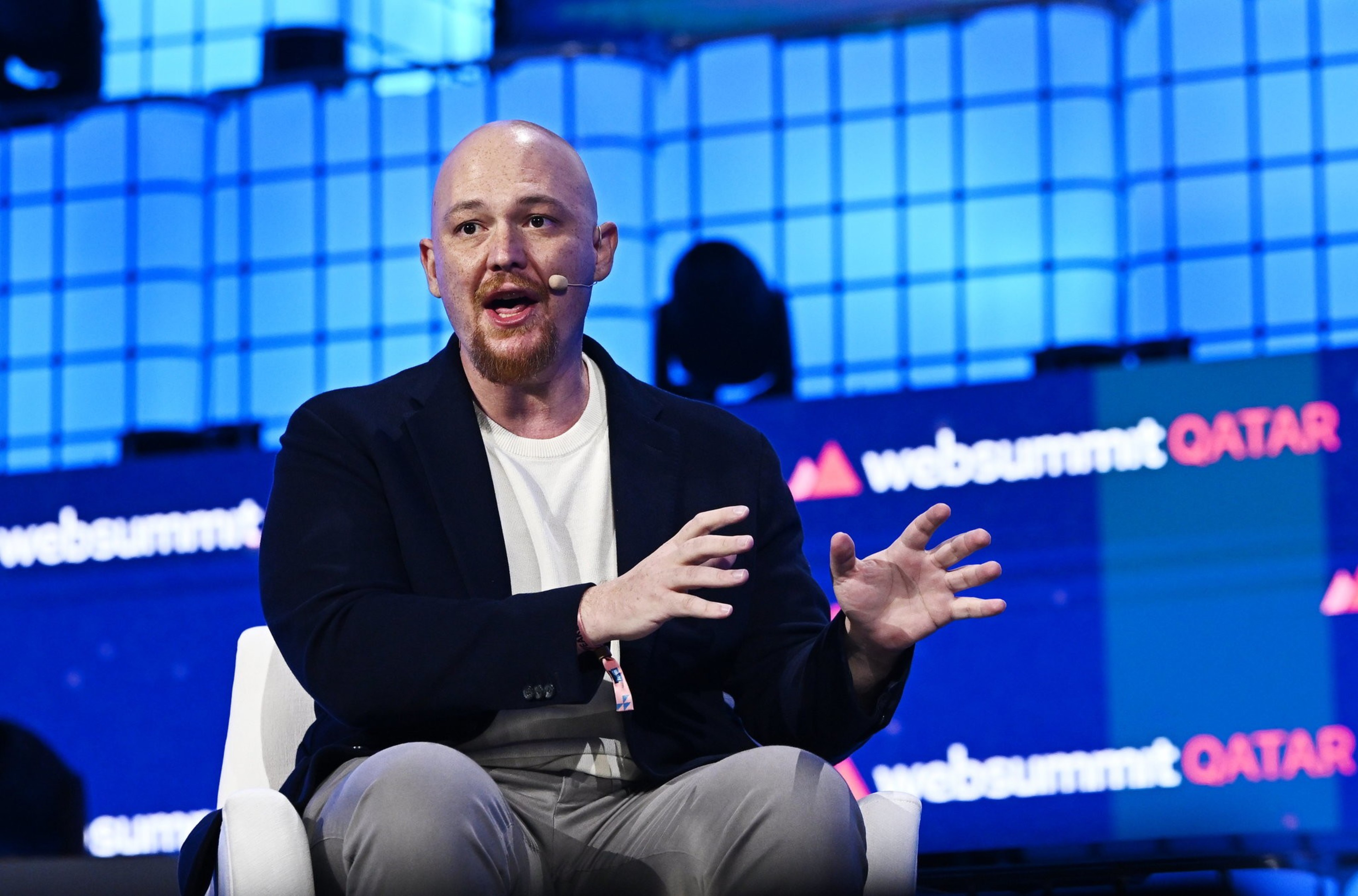 A bald man with a beard and headset mic is seated, gesturing with his hands while speaking at Web Summit Qatar, with the event name displayed on screens behind him.