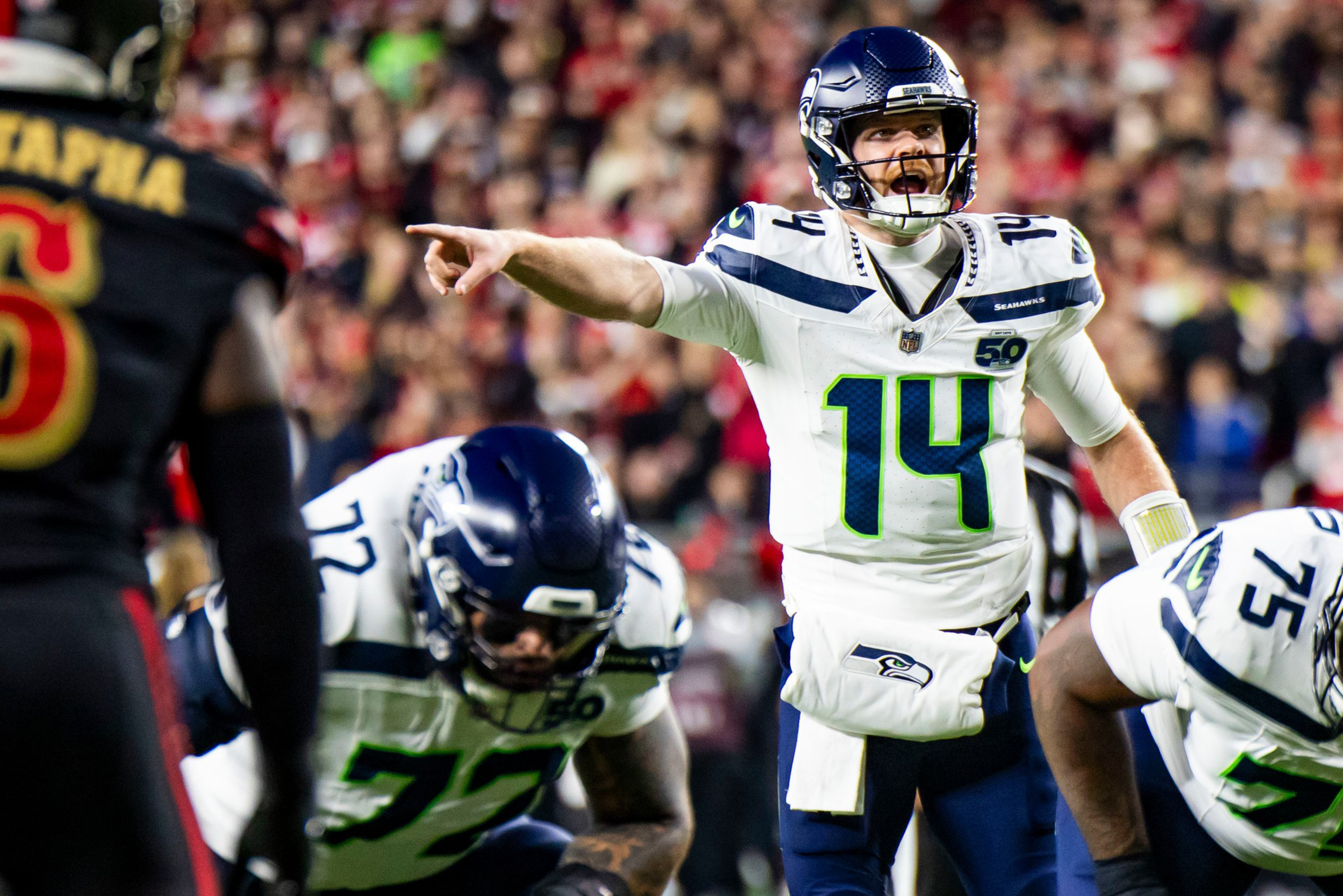 A Seattle Seahawks quarterback wearing number 14 points forward while calling a play, surrounded by teammates and facing a defender in black and red.