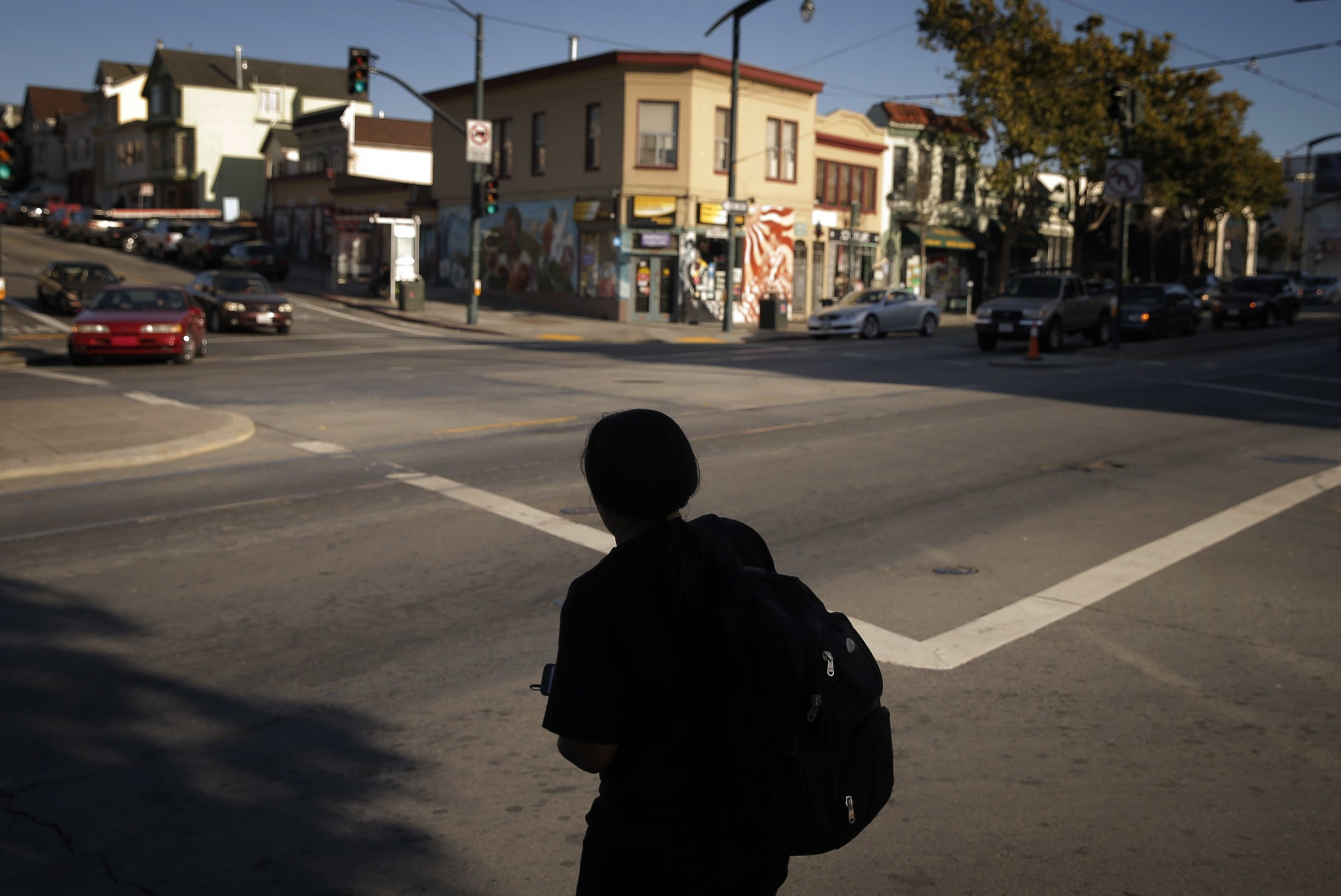 A person with a backpack stands at an empty crosswalk on a city street corner with buildings and cars in the background under late afternoon sunlight.