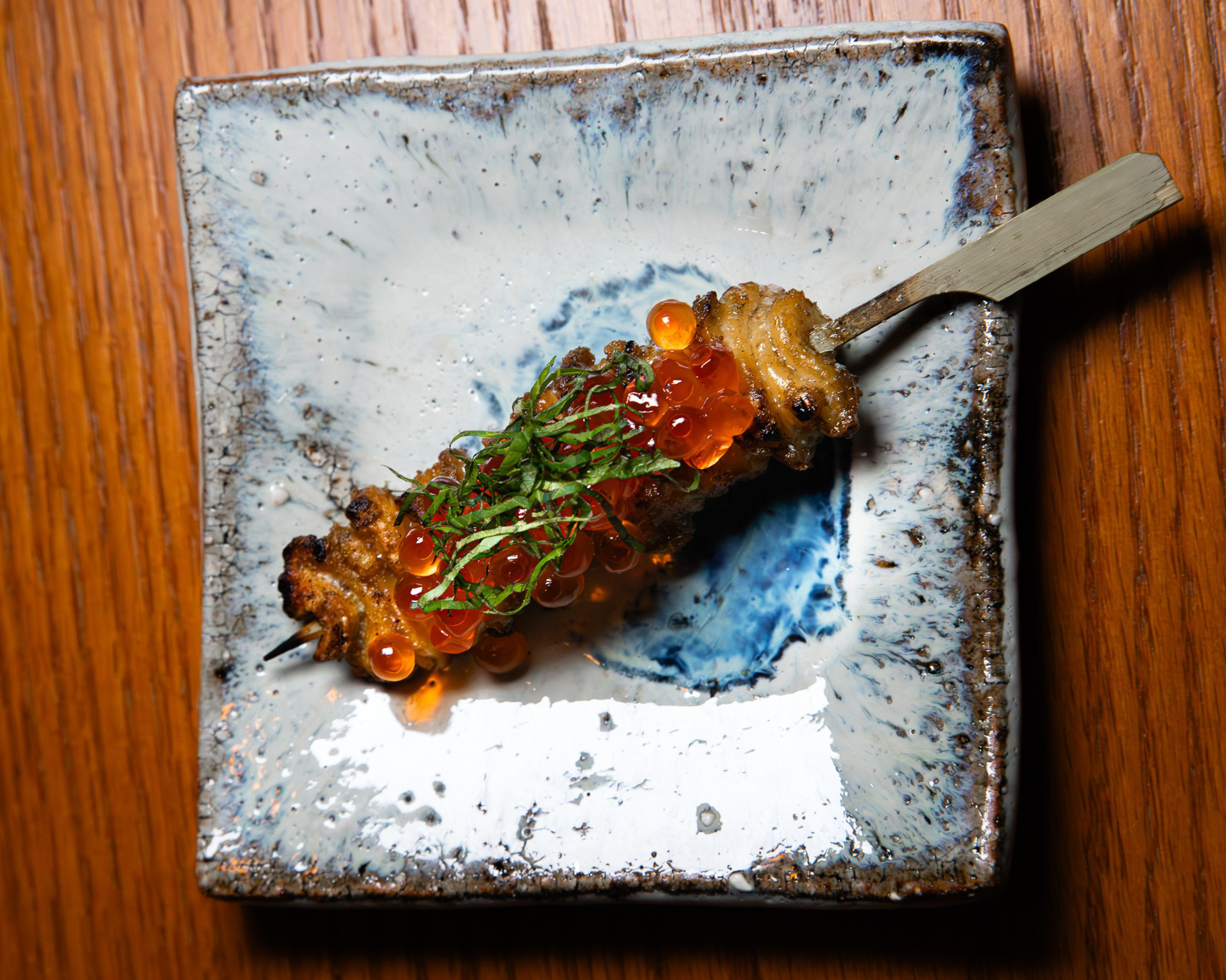 A grilled skewer topped with bright orange salmon roe and thinly sliced green herbs rests on a square ceramic plate with a blue and white glaze.