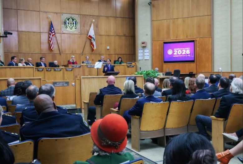 Dozens of people in wood-backed seats on either side of an aisle watch a speech by a man standing behind a dais with several other people seated on a stage behind him. It.