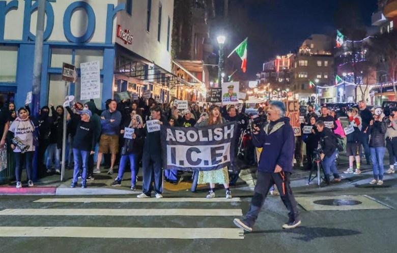 A crowd of people gathers in Little Italy in a crosswalk, holding signs and protesting a fatal shooting by ICE
