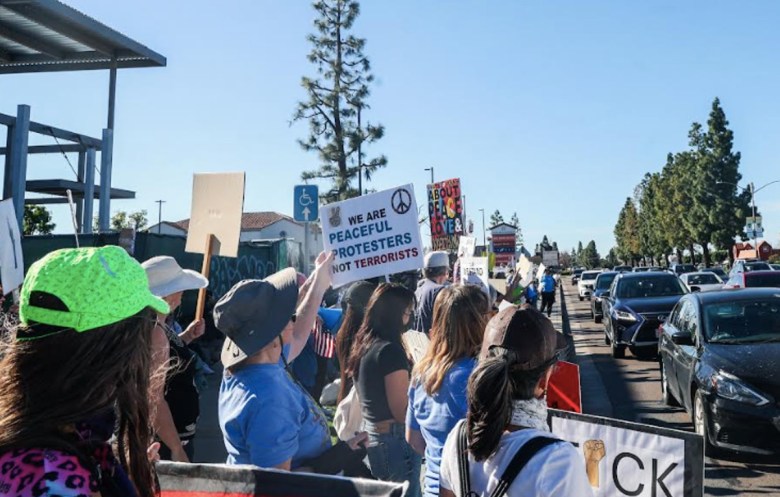 A number of people stand holding signs on a public street with several cars seen in front of them. They are protesting the killing of Renee Good.
