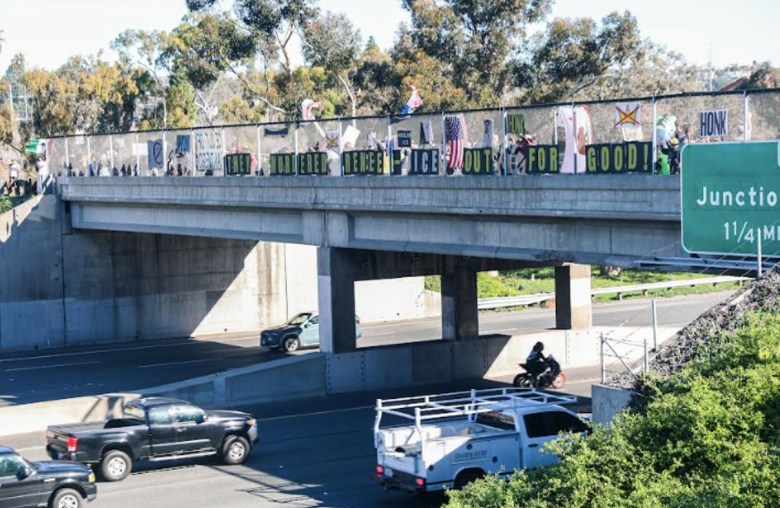 A line of people stand behind a fence on an overpass over a freeway. On the fence are various signs, including a one that reads "They murdered Renee - ICE out for good."