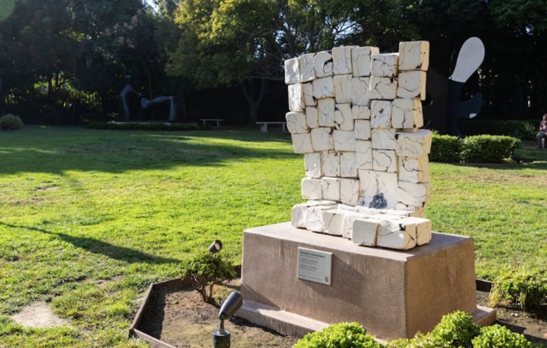 An outdoor sculpture, with what a stack of what could be slips of paper in the shape of a sail on a wooden base in the midst of a garden.