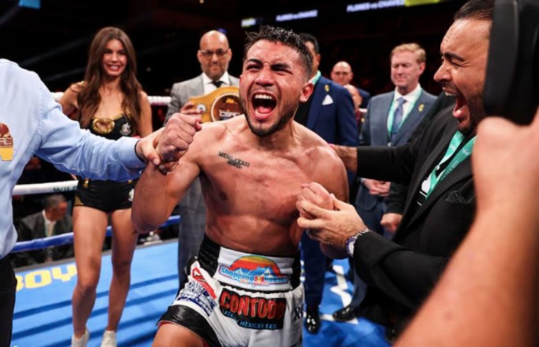 A man in a boxing ring surrounded by several people exults as the referee reaches toward his arm, indicating that he won the bout. The boxer is Jorge “El Niño Dorado” Chavez of San Diego.