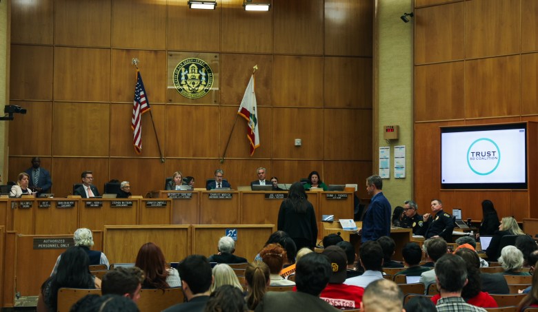 Flags hang at the front of a room over several people sitting and watching over a packed audience. Two people stand speaking before them and a screen on the wall shows a sign "Trust SD Coalition."