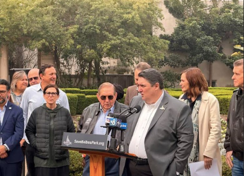About 10 people stand behind a podium set up in a grassy area with trees as a backdrop.