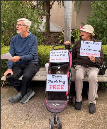A man and a woman sit on a park bench with a stroller between them carrying a dog, a papillon. The hold signs objecting to new parking fees at Balboa Park; one calls the park "our commons."