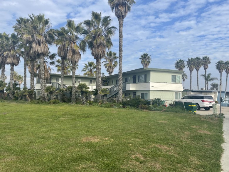 a broad stretch of grass in front of a neatly kept two-story apartment building with palm trees in front and a bright blue sky with sparse clouds as a backdrop.