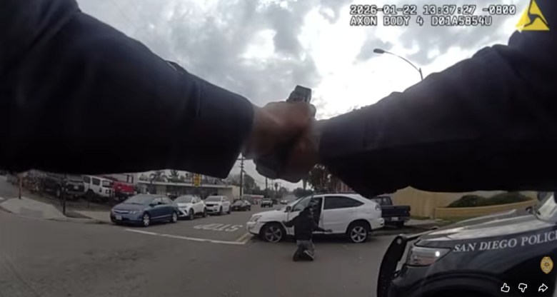 Two arms are extended on a city street with a man kneeling on the ground several feet in front of the person aiming a firearm at him. The man on his knees is standing in front of a compact SUV. 