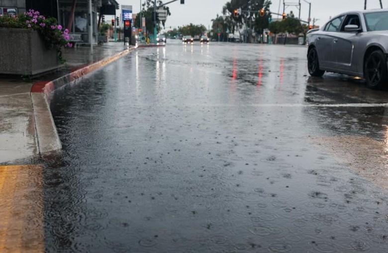 Raindrops can be seen in a puddle near the curb of a city street with a sports car going by.