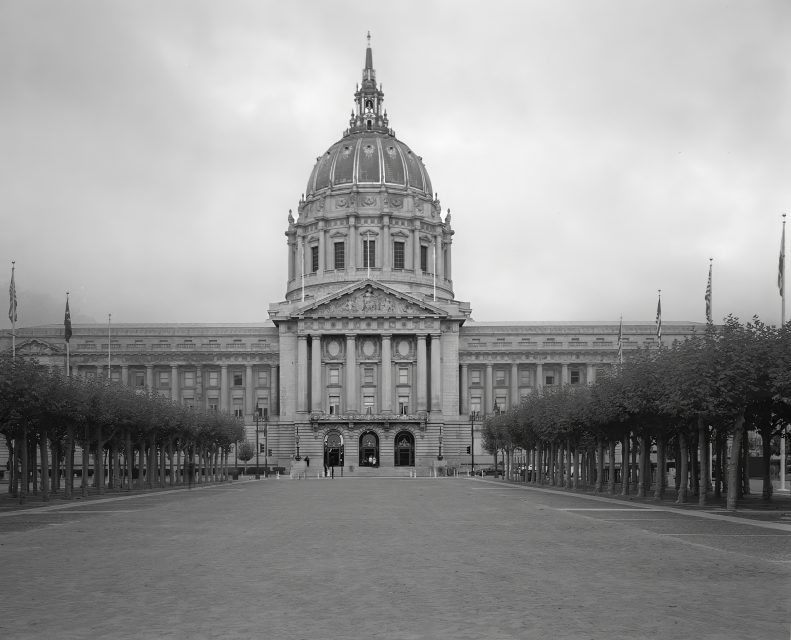 A large neoclassical building with a prominent dome, symmetrical facade, columned entrance, and a wide plaza lined with trimmed trees.
