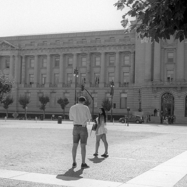 Two people stand in front of a large, historic building with columns on a sunny day; trees and street signs are visible in the background.