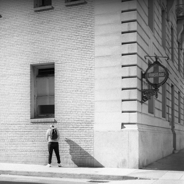A person wearing a backpack stands facing a brick wall next to a building with a "First Aid" sign, in an urban setting.