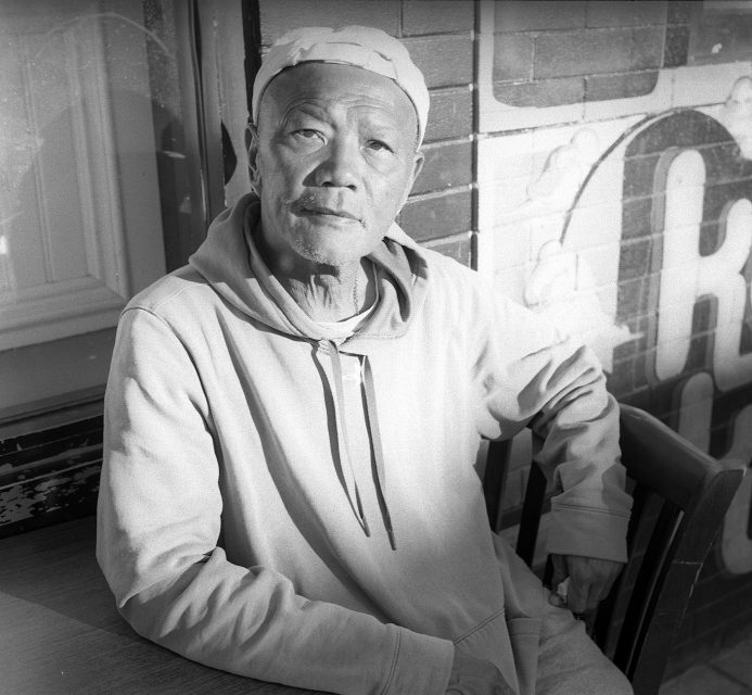 An older man in a hoodie and cap sits at a table by a window, looking at the camera; brick wall with painted lettering in the background.