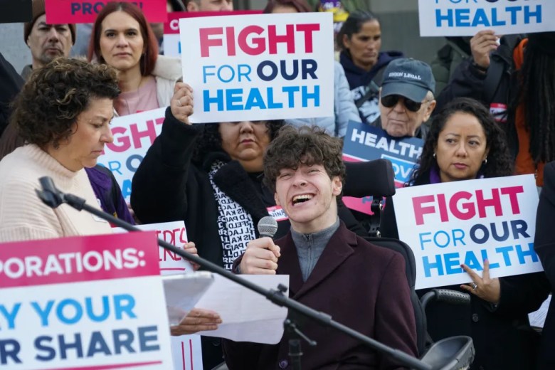 A person using a wheelchair speaks into a microphone at a rally, smiling while holding notes, as people behind them hold signs reading “Fight for Our Health” and “Pay Your Fair Share.”