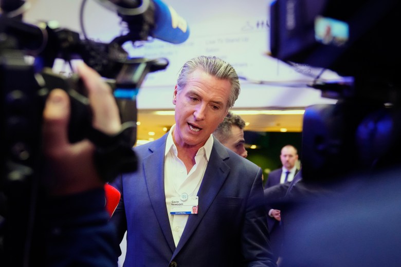 Gov. Gavin Newsom speaks to reporters while surrounded by television cameras and microphones at an indoor event, wearing a dark suit and conference badge as security and attendees stand nearby.