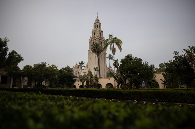 A beige and brown Spanish-style tower is seen through the green bushes of a nearby garden.