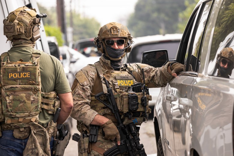 An immigration agent, dressed in camouflage riot gear and holding a weapon, looks straight forward while standing next to a car as their right arm rests on the driver's window seat.