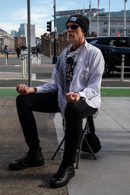A man wearing sunglasses, a beanie, and a white shirt sits cross-legged on a folding stool on a city sidewalk, holding his hands in a meditative pose.