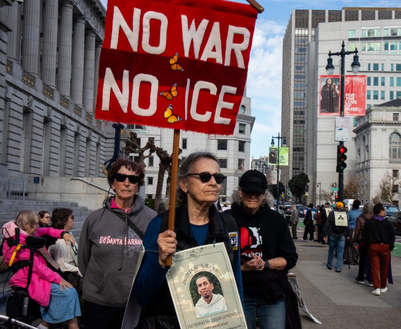 A woman at a protest holds a red sign reading "No War No ICE" and a photo of a person; others stand behind her on a city street near government buildings.