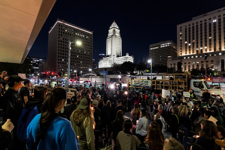 A large crowd gathers at night in a downtown plaza, many holding candles and protest signs, with city hall and office buildings lit in the background as traffic passes along a busy street.