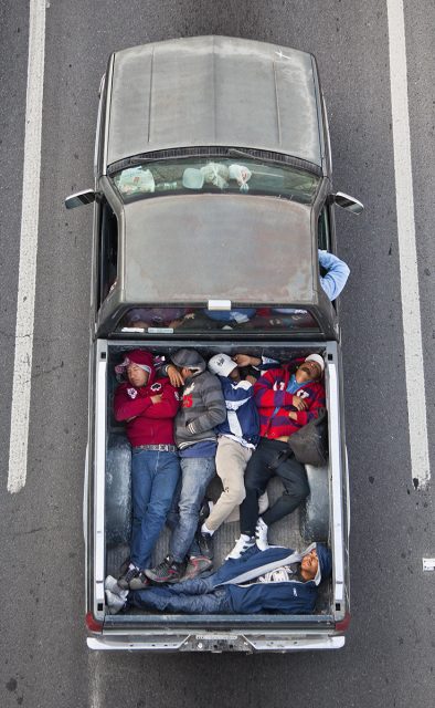 Aerial view of six people lying closely together in the bed of a pickup truck parked between two white lines on a road.