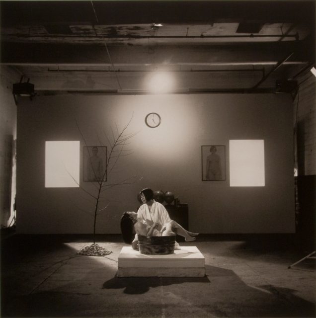 A person in a white robe holds another person in a basin on a platform in a dimly lit room with a bare tree, a clock, and two portraits on the wall.
