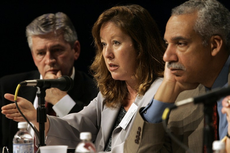 A person with strawberry blonde hair sits in between two other people as they gesture in front of them while speaking in front of a group of people.
