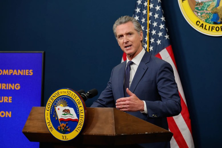 A person in a suit speaks at a podium with the California state seal, flanked by the U.S. flag. A screen displaying text is on their left.