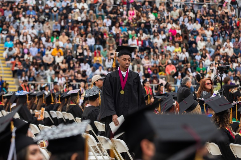 A graduate in a black cap and gown walks through rows of seated graduates holding a diploma, with a large crowd filling the stadium stands in the background