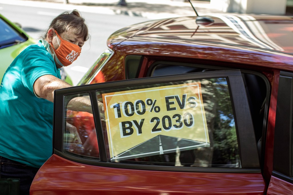 A person wearing a face mask places a sign reading “100% EVs by 2030” inside the open rear door of a parked car on a tree-lined street.