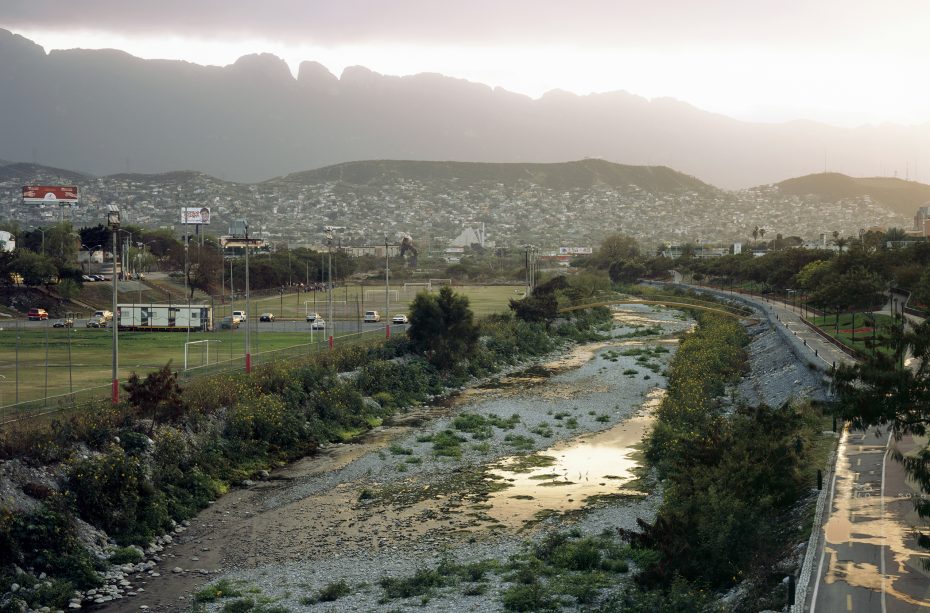 A shallow, rocky river flows through an urban area with cars on a nearby road, soccer fields, and mountains in the background at sunset.
