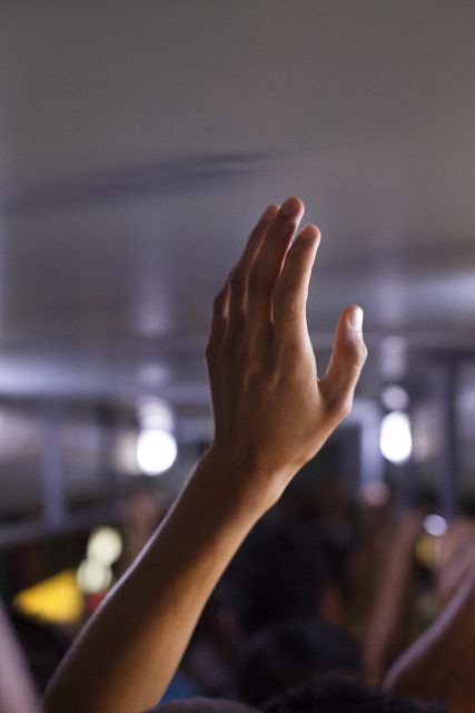 A raised hand in focus inside a crowded, dimly lit space, possibly a bus or train, with blurred people in the background.