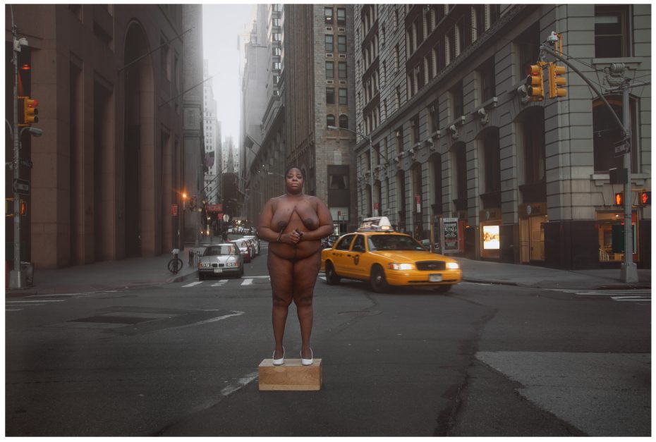 A person stands nude except for white shoes on a wooden box in the middle of a city street, surrounded by tall buildings and traffic, including a yellow taxi.