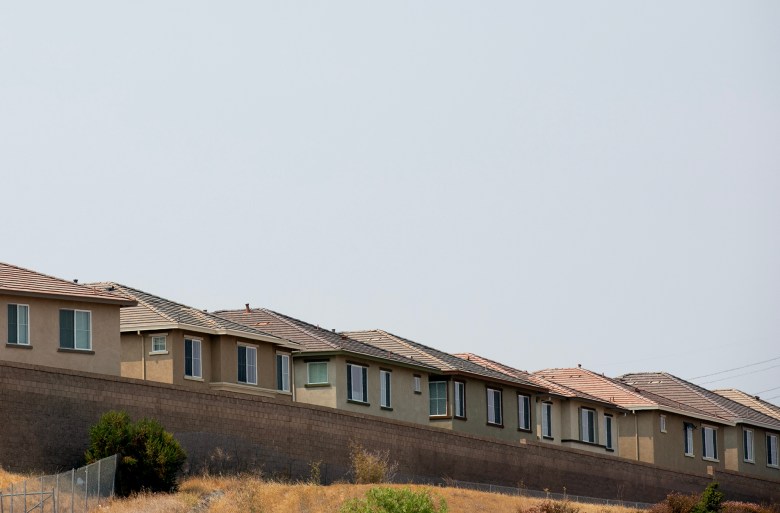 A row of beige suburban houses with tiled roofs lines the top of a sloped retaining wall, with dry grass and sparse shrubs in the foreground beneath a wide, pale sky.