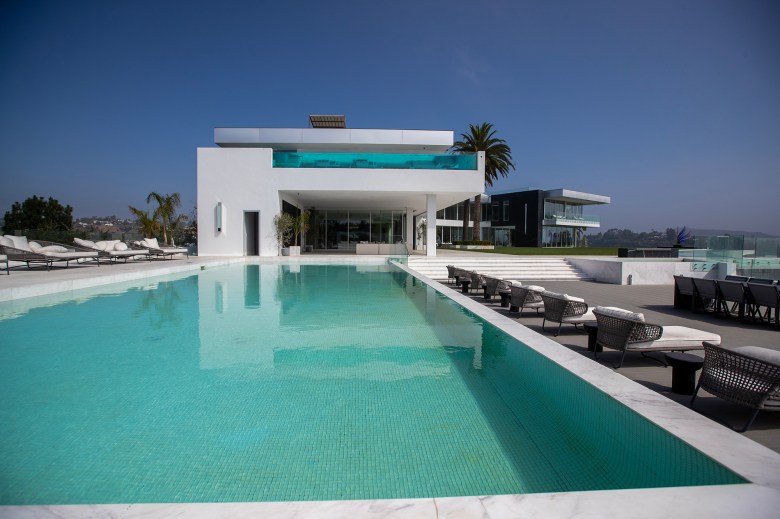 A modern white hillside mansion features a long turquoise swimming pool in the foreground, with lounge chairs lining the deck and expansive glass walls opening to outdoor living areas under a clear blue sky.