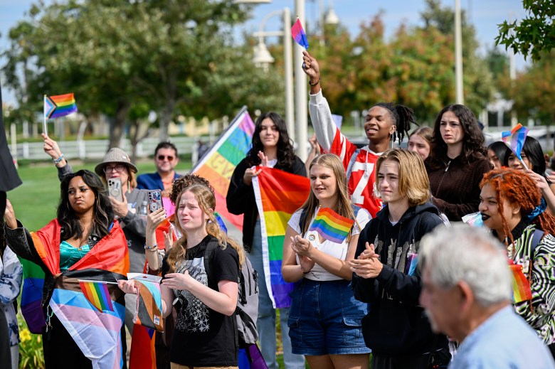 A diverse group of people gathers outdoors, holding and waving various LGBTQ+ pride flags, including the progress pride flag, rainbow flag, and bisexual pride flag. Many attendees are clapping, smiling, and recording the event on their phones. Some individuals are draped in flags, while others hold them high. The background features trees, lampposts, and a grassy area, suggesting a public park or school campus setting.